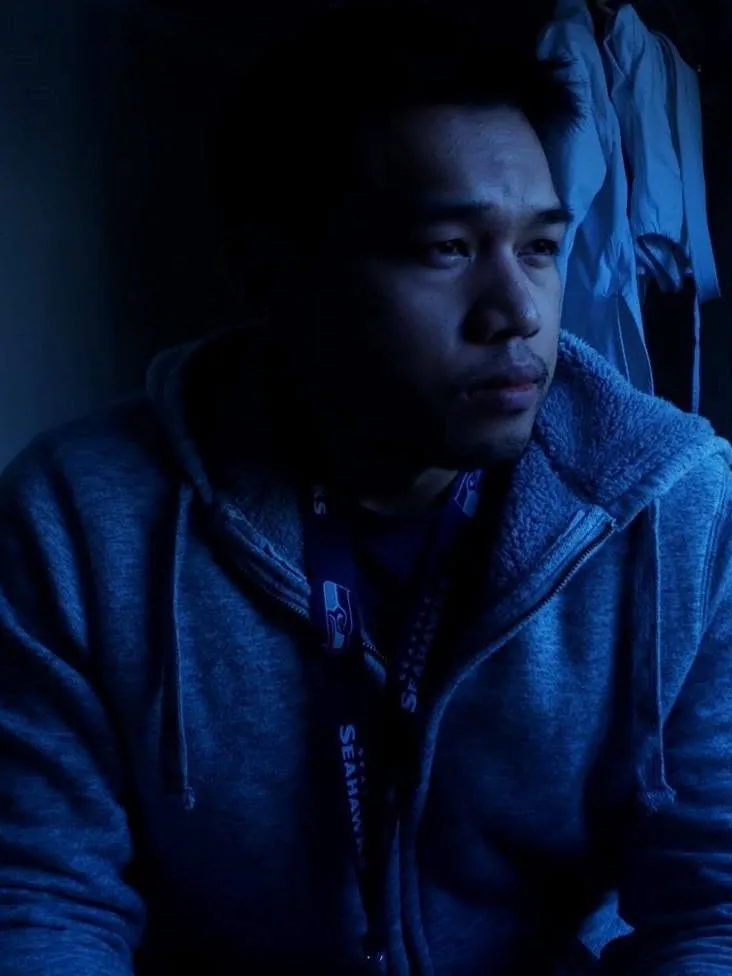 Ermenildo Castro, a real photograph: a man in a gray hoodie and lanyard, in a dim blue room.